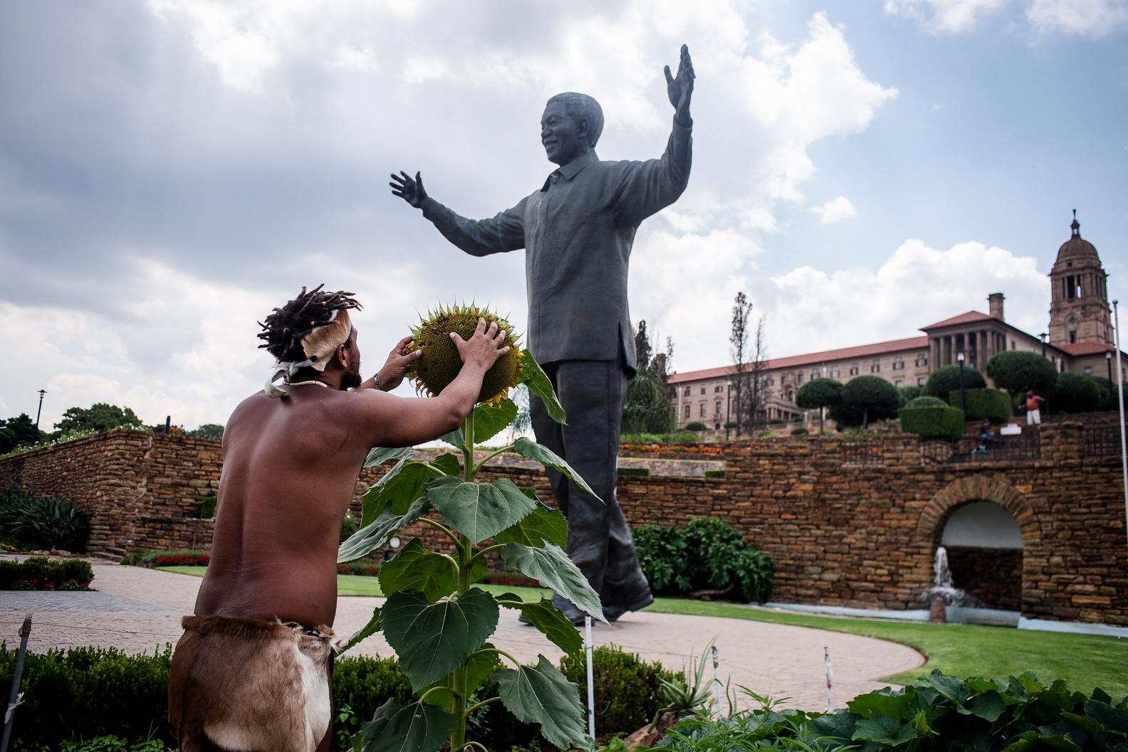 A Khoisan man stands with both arms outstretched holding a sun flower, terraced gardens and a government building on the hill behind.